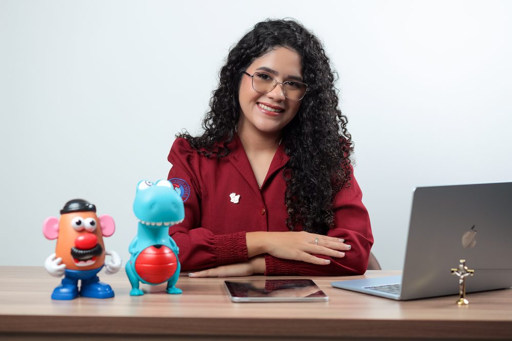 Gabrielle Aquino sentada à mesa de trabalho no Vitória Instituto Terapêutico, com brinquedos usados em terapia infantil, tablet e notebook.