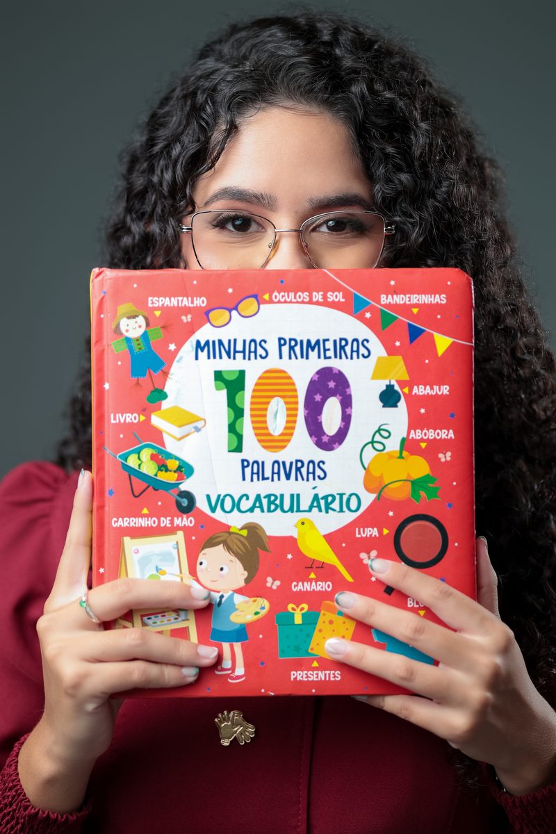 Gabrielle Aquino segurando um livro infantil de vocabulário à frente do rosto, de forma lúdica.