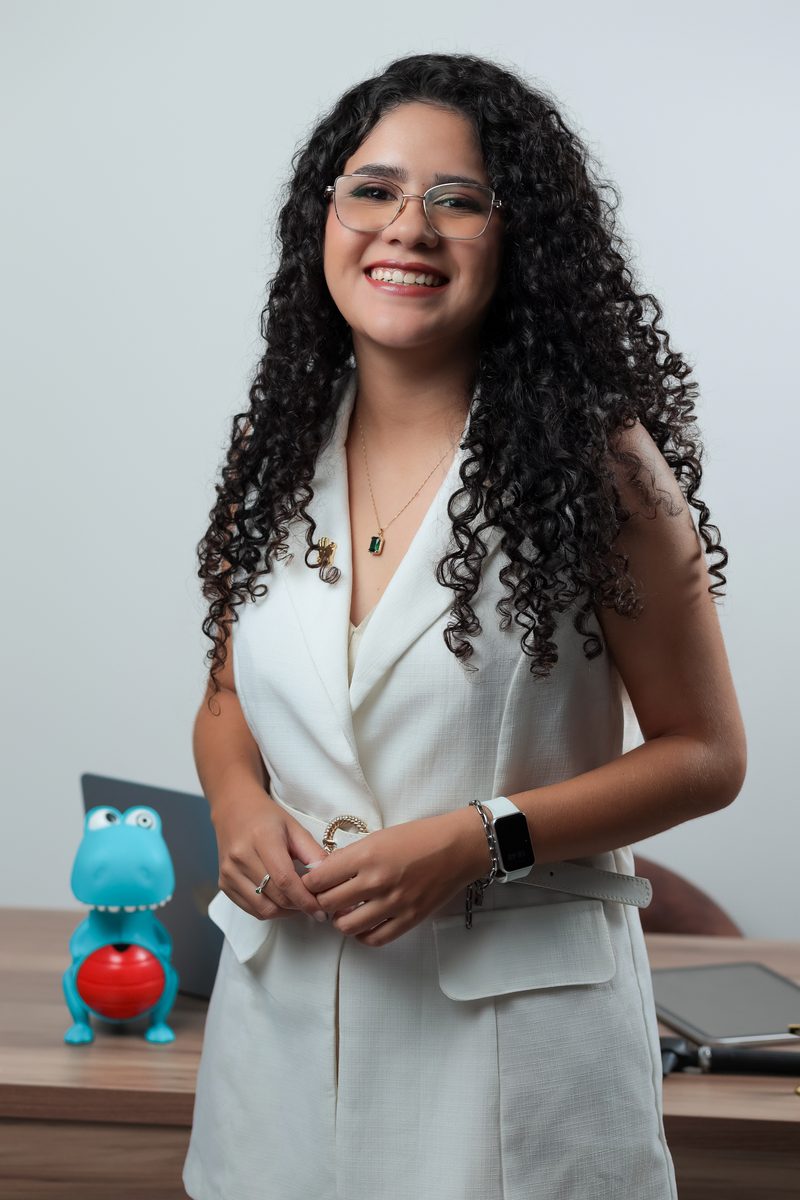 Gabrielle Aquino em pé junto à mesa de trabalho, com um brinquedo de dinossauro e notebook ao fundo.