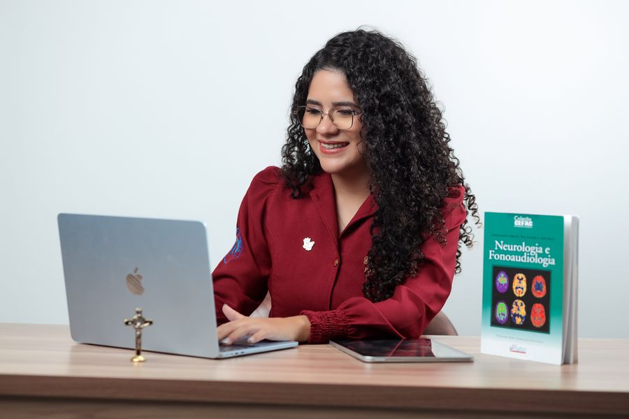Gabrielle Aquino trabalhando ao notebook, com um livro de Neurologia e Fonoaudiologia ao lado, em sua mesa de trabalho.