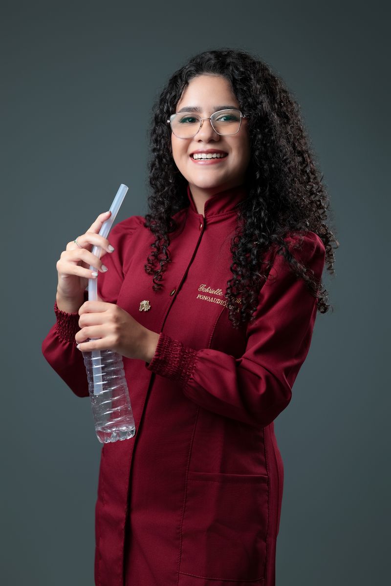 Gabrielle Aquino, com o jaleco vinho do consultório, segurando um canudo de ressonância usado em terapia vocal.